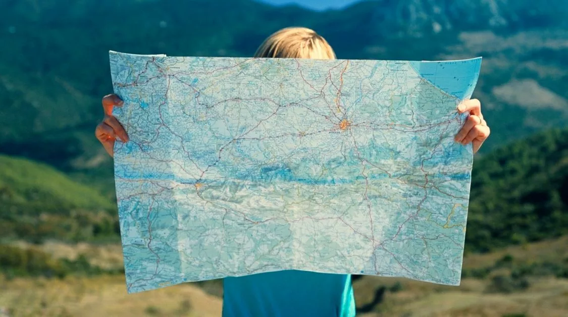 planning ahead for the tax year-end: image shows a person hidden behind a large map, looking at direction. They are outside, with hills in the distance behind them.