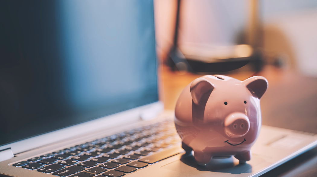 Lifetime Allowance is to be abolished from 6 April 2024. Image shows a pink, smiling piggy bank sitting on a grey laptop, with a warm, blurred out background.