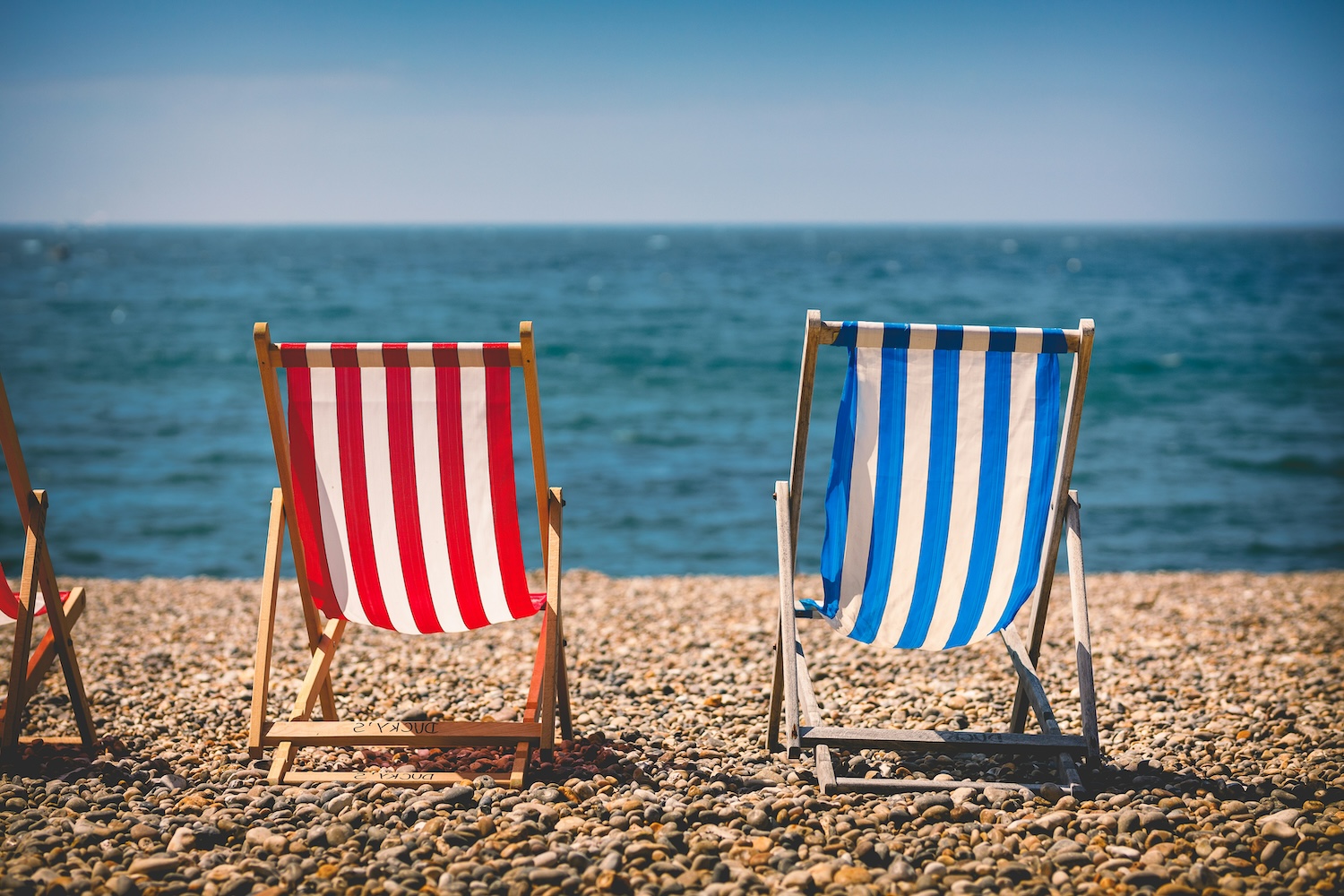 Can I afford to retire? Image depicts two sun loungers on a pepple beach somewhere in the UK. One chair is red and white striped, the other blue and white. It is a sunny day, and both chairs are facing out towards the sea.